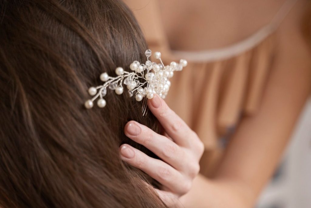 Aide pour savoir quel accessoire cheveux choisir pour la mariée : pose d'un peigne délicat en perles blanches et cristaux dans des cheveux bruns ondulés.